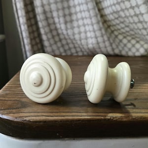May include: Two off-white, vintage-style cabinet knobs with a circular design. One knob is lying on its side, while the other is upright. The knobs are set against a wooden surface.