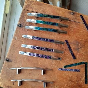 May include: A collection of cabinet hardware, including drawer pulls and handles, displayed on a wooden surface. The pulls feature silver metal with colorful, speckled inlays of green, blue, and purple. Some handles have a simple, curved design.