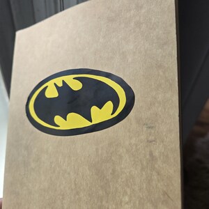 Pode incluir: Um caderno retangular com uma capa de papel texturizado castanho-claro. Centrado na capa frontal est&aacute; um gr&aacute;fico do log&oacute;tipo do Batman, apresentando uma silhueta de morcego preta sobre um fundo oval amarelo brilhante.