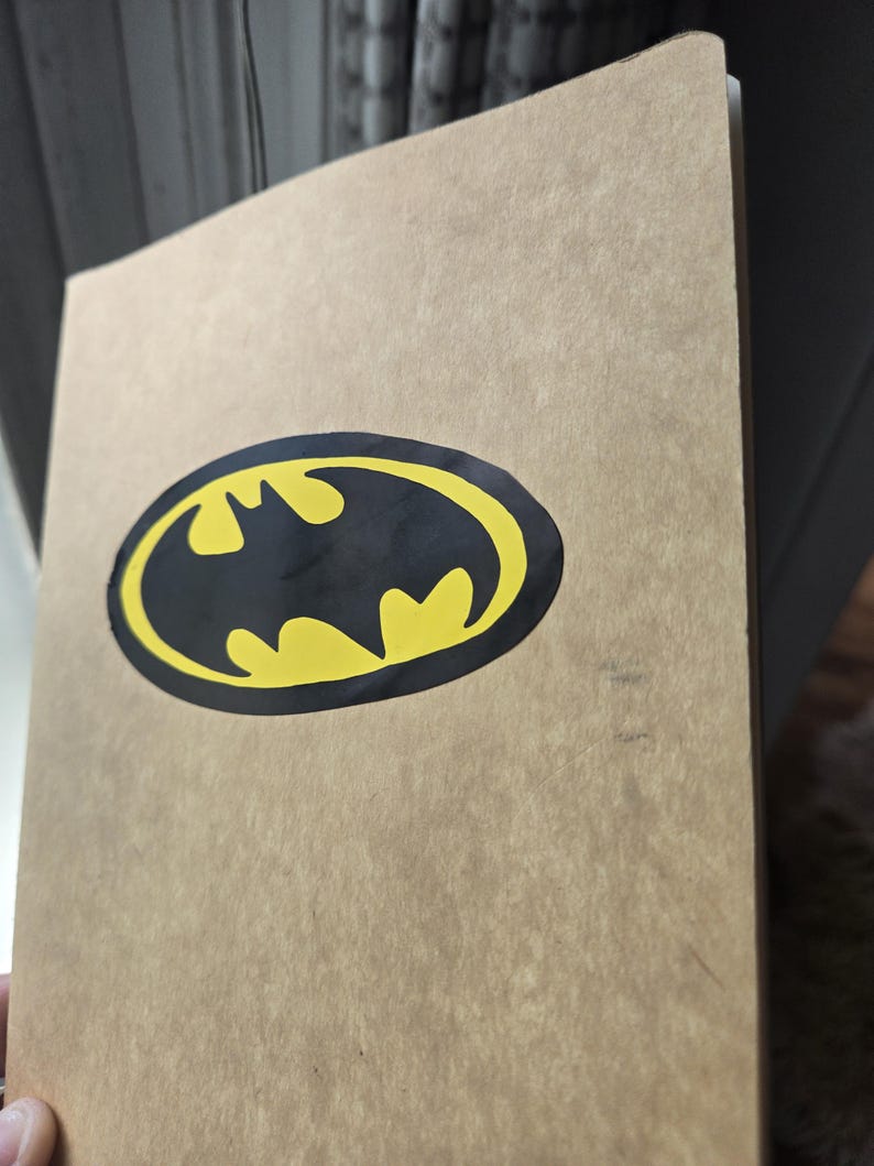 Pode incluir: Um caderno retangular com uma capa de papel texturizado castanho-claro. Centrado na capa frontal est&aacute; um gr&aacute;fico do log&oacute;tipo do Batman, apresentando uma silhueta de morcego preta sobre um fundo oval amarelo brilhante.