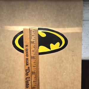 Pode incluir: Um autocolante com o log&oacute;tipo do Batman em preto e amarelo sobre uma superf&iacute;cie bege, colocado junto a uma r&eacute;gua de madeira que mostra uma altura de cerca de 6,35 cm. A r&eacute;gua apresenta o texto A GOOD RULE Do Unto.