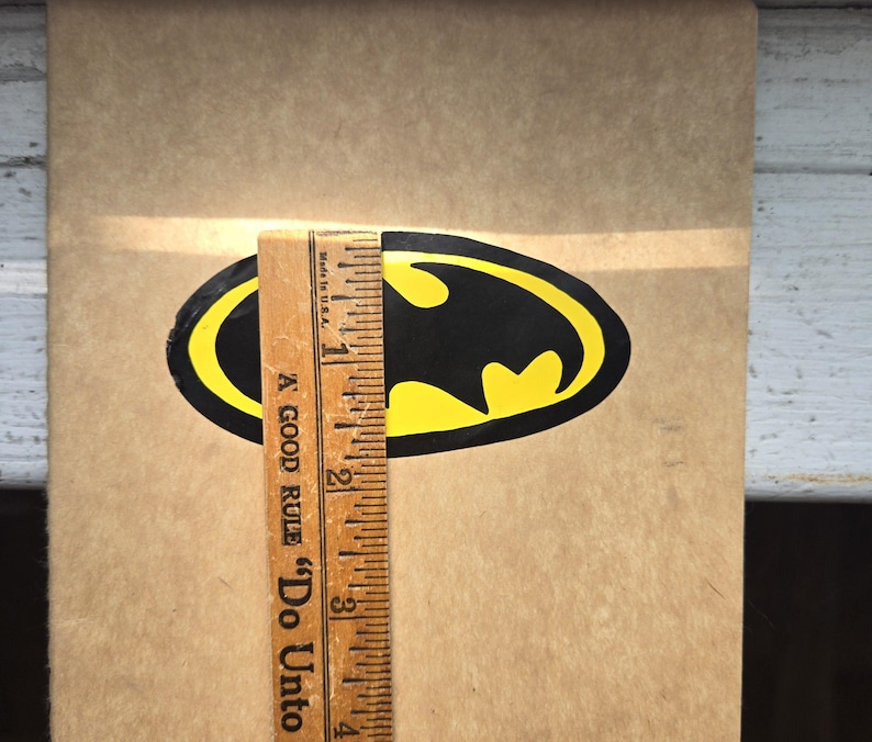 Pode incluir: Um autocolante com o log&oacute;tipo do Batman em preto e amarelo sobre uma superf&iacute;cie bege, colocado junto a uma r&eacute;gua de madeira que mostra uma altura de cerca de 6,35 cm. A r&eacute;gua apresenta o texto A GOOD RULE Do Unto.