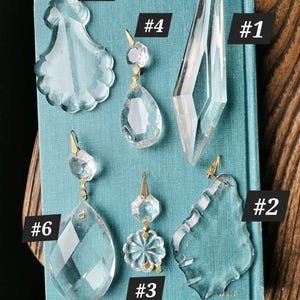 May include: Six clear glass chandelier crystals labeled #1 through #6. The collection includes various shapes: a long diamond, two scalloped teardrops, two faceted teardrops with brass connectors, and one small circular faceted piece with a brass connector.