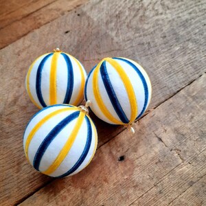 May include: Three white Christmas ornaments with blue and yellow stripes. The ornaments are made of fabric and have a velvet texture.