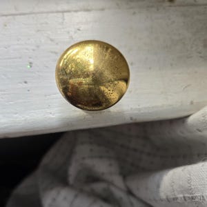 May include: A round, brass-colored cabinet knob with a slightly textured surface. The knob is mounted on a white painted surface, with a glimpse of a white fabric in the background. The knob has a vintage appearance.