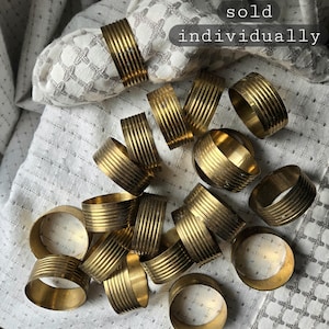 May include: A collection of gold-colored metal napkin rings with a ribbed design. The rings are scattered on a white and gray patterned cloth. The text "sold individually" is visible.