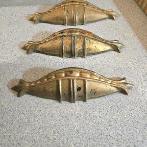May include: Three antique gold-toned cabinet drawer pulls with a decorative design. The pulls are in the shape of a shell with a raised center and a scalloped edge.
