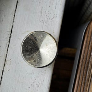 May include: A circular, metallic cabinet knob with a polished, reflective silver finish. The surface features a subtle, conical indentation that tapers toward the center, creating a radial light pattern across the smooth, circular face of the hardware.