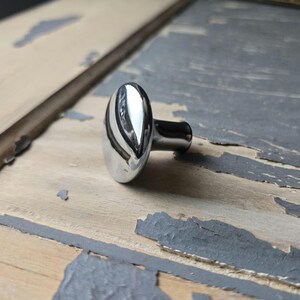 May include: A polished chrome cabinet knob with a smooth, rounded, oval-shaped face and a cylindrical base. The metallic surface is highly reflective, showing light highlights and a curved silhouette that tapers toward the mounting point.