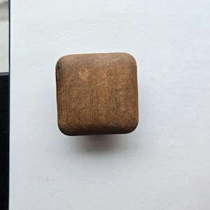 May include: A small, rectangular wooden block with rounded edges. The block is a medium brown color with visible wood grain and a slightly worn appearance. The object is set against a white background.