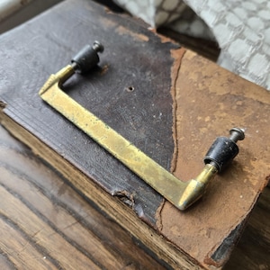 May include: An antique brass drawer pull with black hardware is attached to a worn, leather-bound book. The book's pages are aged, and the leather has a distressed, brown appearance. The pull is rectangular with a right-angle bend.