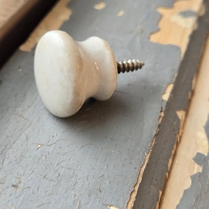 May include: A white ceramic cabinet knob with a smooth, rounded face and a tapered neck. A metal screw with a threaded shank extends from the back of the knob, designed for attachment to furniture or cabinetry surfaces.