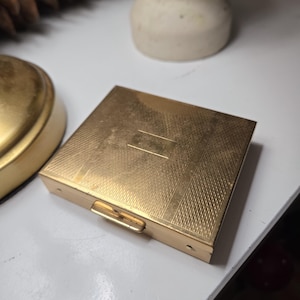 May include: A gold-colored metal compact with a textured surface and a hinged lid. The compact is closed and has a small latch on the side.
