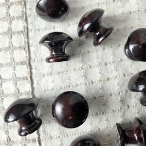 May include: A collection of dark brown wooden knobs in various shapes, including round and mushroom-shaped designs. The knobs are arranged on a white, textured fabric, showcasing their polished finish and potential for furniture or cabinet hardware.
