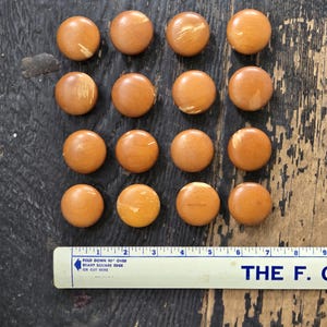 May include: Sixteen round, wooden buttons in varying shades of brown and tan are arranged on a dark wooden surface. A ruler with text is at the bottom. The buttons have a smooth, polished finish and are likely for sewing or crafts.