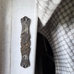 May include: A weathered, bronze-colored metal door hinge with an ornate, textured design. The hinge is mounted on a white painted wooden door frame. The design includes decorative flourishes at the top and bottom, with a central section featuring a textured surface.