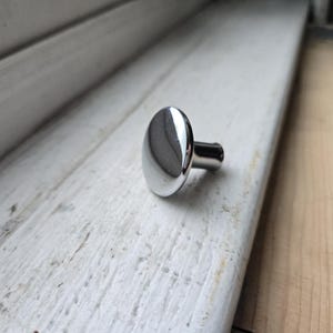 May include: A shiny, chrome-finished oval cabinet knob. The knob has a smooth, reflective surface and a short, cylindrical stem. It is set against a weathered, white-painted wooden surface, suggesting a vintage or rustic aesthetic. The knob is approximately 1.5 inches long.