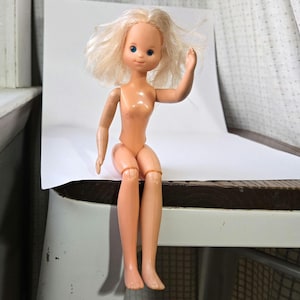 May include: A vintage plastic doll with blonde hair and blue eyes. The doll is wearing a nude-colored outfit and is sitting on a wooden surface.