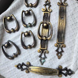 May include: A collection of antique-style brass furniture hardware including five teardrop-shaped swinging drawer pulls and two elongated backplates with ornate scrollwork details and textured centers, all featuring a dark aged patina finish.