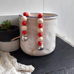 May include: Two strands of wooden beads with alternating red and white beads. Each strand has a silver heart-shaped bell at the end. The beads are hanging from the edge of a white ceramic container.