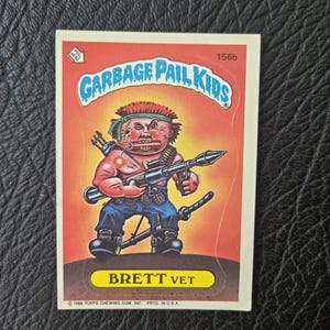May include: A vintage Garbage Pail Kids trading card featuring "Brett Vet." The card has a red background with the Garbage Pail Kids logo at the top. The illustration depicts a cartoon character holding a weapon and wearing a headband.
