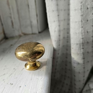 May include: A close-up of a gold-colored cabinet knob with an oval shape. The knob has a textured surface and a small, round base. The knob is set against a white surface and a white and gray patterned curtain.