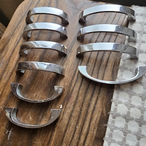 May include: A collection of silver-toned cabinet handles arranged on a wooden surface. The handles have a curved, arched design, and some show signs of wear. The background includes a patterned fabric and a wooden surface.