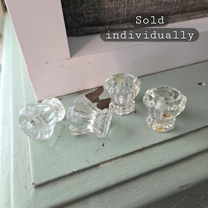 May include: Four clear glass cabinet knobs with a faceted design. The knobs are displayed on a light blue painted surface. The text "Sold individually" is visible in the image.
