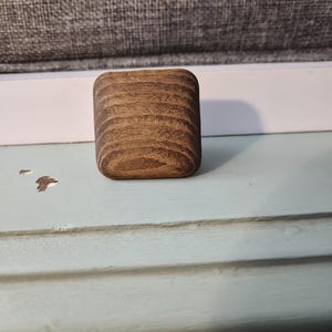 May include: A small, rectangular wooden block with rounded edges. The wood grain is visible, showcasing a warm brown color with darker streaks. The block is sitting on a light blue surface.