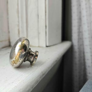 Pode incluir: Um puxador de armário prateado em forma de cogumelo com um tom dourado manchado. O puxador está colocado sobre uma superfície pintada de branco, sugerindo uma estética vintage ou antiga. O hardware é provavelmente para móveis ou armários.