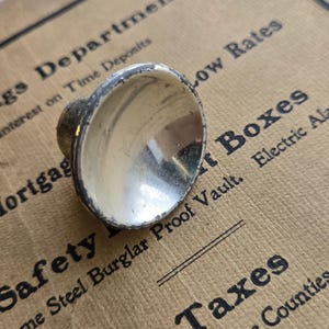 May include: A small, silver-colored knob with a rounded, concave top and a short, cylindrical base. The knob has a slightly worn appearance, with some discoloration around the edges. The background features text related to banking and finance.