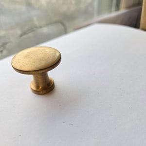 May include: A close-up of a brass cabinet knob with a rounded top and a short, cylindrical base. The knob has a warm, golden hue and a slightly textured surface, suggesting an aged or vintage appearance. The knob is set against a white surface.