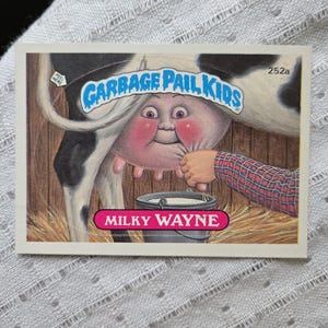 May include: A Garbage Pail Kids trading card featuring a cartoon character with a pink face and a cow's udder. The character is being milked into a bucket. The card is labeled "Milky Wayne."
