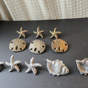 May include: Collection of decorative metal coastal-themed objects, including starfish, sand dollars, and conch shells. The objects are in various shades of silver and gold, arranged on a dark surface.