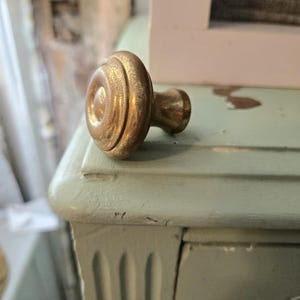 May include: A gold-colored, antique-style cabinet knob with a circular design. The knob is resting on a light blue painted wooden surface. The knob has a round face with concentric circles and a short, cylindrical stem.