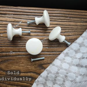 May include: Four round, off-white ceramic cabinet knobs with smooth, glossy finishes. Each knob features a central metal screw attachment. The text 'Sold individually' is visible in the lower left corner of the image, indicating purchase options.