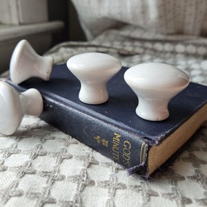 May include: Three white ceramic cabinet knobs with a smooth, rounded top and a tapered base. The knobs are positioned on top of a dark blue book with the gold text "GOD'S MINUTE" printed on the spine, featuring a small gold floral emblem.