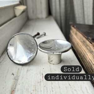May include: Two vintage, silver-toned cabinet knobs with a round, reflective surface and a short, cylindrical base. The knobs are on a white, weathered windowsill, with an antique book visible in the background. The text "Sold individually" is also visible.
