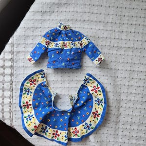 May include: A blue doll outfit featuring a long-sleeved top and a skirt. The outfit is decorated with a floral pattern in red, yellow, and blue. The top has a high collar and the skirt is a circular shape.