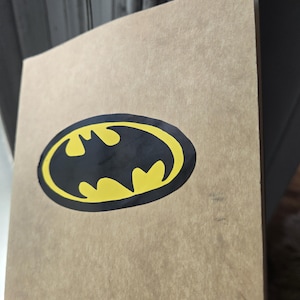 Pode incluir: Um caderno retangular com uma capa de papel texturizado na cor bege. Centralizado na capa frontal, h&aacute; um emblema oval preto com uma silhueta de morcego amarela, representando o ic&ocirc;nico logotipo do Batman.