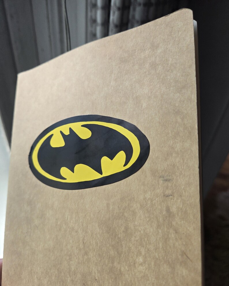 Pode incluir: Um caderno retangular com uma capa de papel texturizado na cor bege. Centralizado na capa frontal, h&aacute; um emblema oval preto com uma silhueta de morcego amarela, representando o ic&ocirc;nico logotipo do Batman.