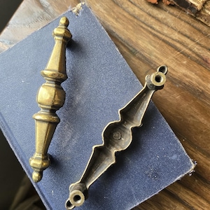 May include: Two antique brass drawer pulls. One is a vertical, ornate handle with a bulbous center and a pointed top. The other is a horizontal pull with a decorative, scalloped design. Both are set against a blue book on a wooden surface.