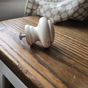 May include: A light-colored wooden cabinet knob with a screw, resting on a wooden surface. The knob has a rounded shape with a decorative design. The wood grain of the surface is visible.