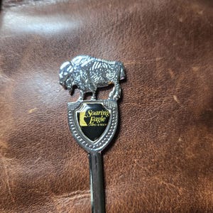 May include: A silver-toned metal souvenir spoon featuring a sculpted buffalo figure standing atop a shield-shaped emblem. The black shield displays the yellow text Soaring Eagle Casino & Resort. The spoon has a polished finish and a long handle.
