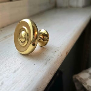 May include: A shiny, gold-colored cabinet knob with a circular design. The knob is resting on a white painted wooden surface. The knob's design includes concentric circles and a small, rounded center.