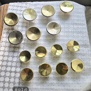 May include: A collection of round, brass-colored cabinet knobs. The knobs are arranged on a white, textured fabric. The image includes the text "sold individually".