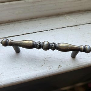 May include: Antique brass cabinet pull with a decorative design. The handle features a central beaded section flanked by rope-like detailing and rounded ends. The pull is mounted on two gray metal posts.