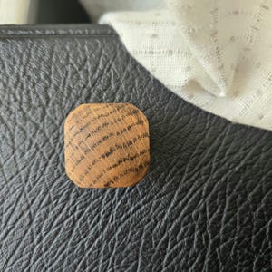 May include: A small, square wooden object with rounded corners. The wood has a natural, light brown color with dark brown grain lines. The object is resting on a textured, black surface, with a white, patterned fabric in the background.