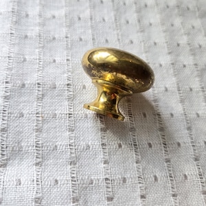 May include: A vintage brass cabinet knob with an oval head and a short, decorative base. The knob has a worn, aged appearance, suggesting it is an antique or a reproduction of an antique. The knob is likely used for furniture restoration or as a decorative element.