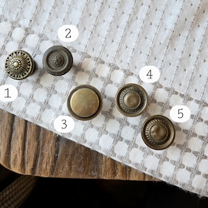 May include: An assortment of five antique brass cabinet knobs. The knobs feature various designs, including floral, circular, and embossed patterns. The knobs are arranged on a white patterned cloth, showcasing their vintage aesthetic.